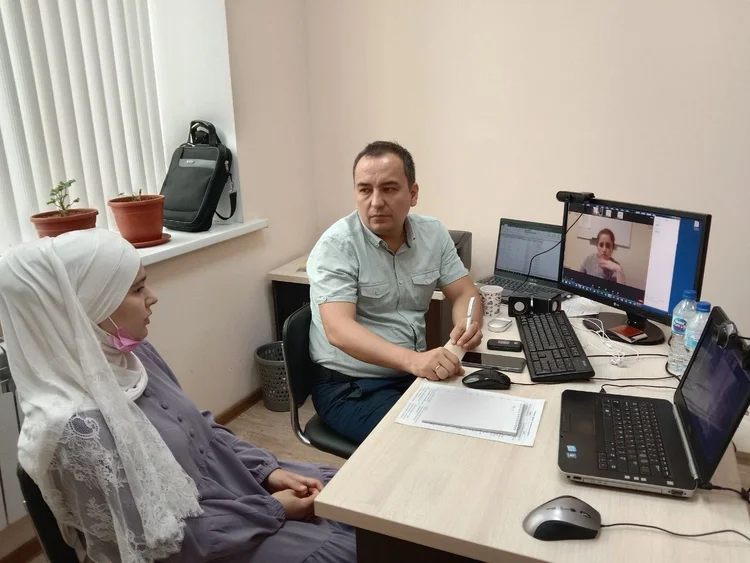 A Uzbek beekeeper and her translator sit at a desk; Sarah's face is visible on the monitor during a Zoom interview